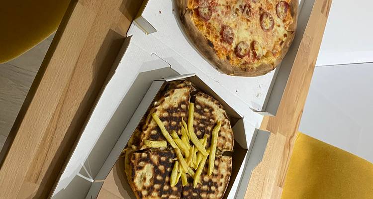 Pizza para llevar y pan plano con papas fritas servidos en cajas de cartón sobre una mesa de cocina.