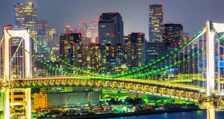 Die grün beleuchtete Rainbow Bridge spannt sich über die Bucht von Tokio vor einer glitzernden Wolkenkratzerskyline in der Nacht.