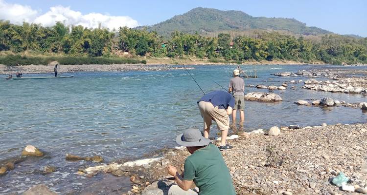 Des voyageurs debout et accroupis sur une berge rocheuse casting des lignes de pêche dans une large rivière turquoise avec des collines forestières au-delà.