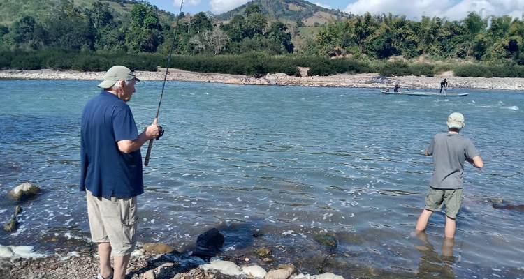 Deux hommes pêchant le long d'une rivière bleue aux eaux vives avec des montagnes et une forêt de bambous en arrière-plan.
