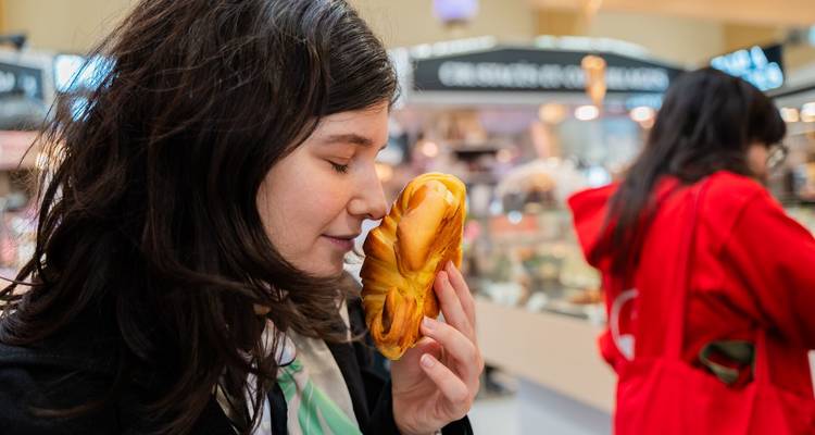 Una mujer joven inhala el aroma de un pastel recién hecho dentro de un mercado cubierto lleno de vida.