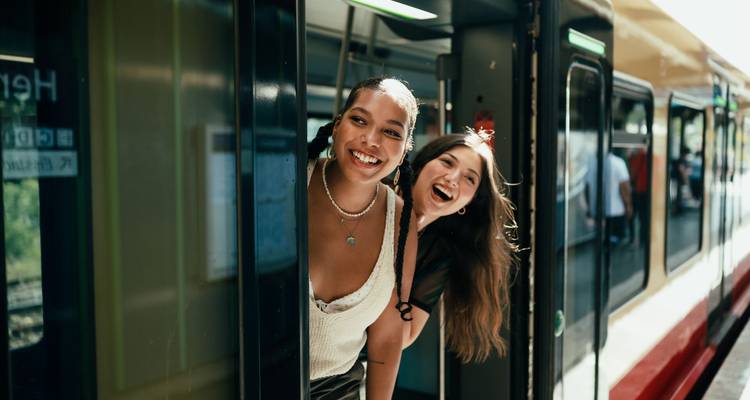 Dos viajeros sonrientes se asoman por la puerta del tren disfrutando del recorrido por el campo.