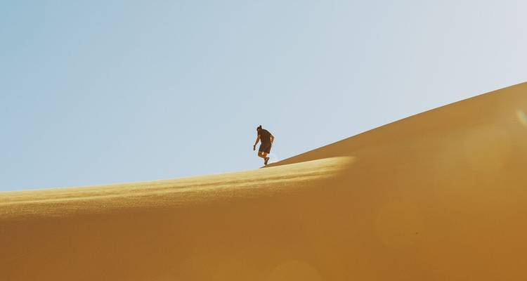 Un aventurier solitaire marche au sommet d'une imposante dune de sable doré sous un ciel bleu dégagé.
