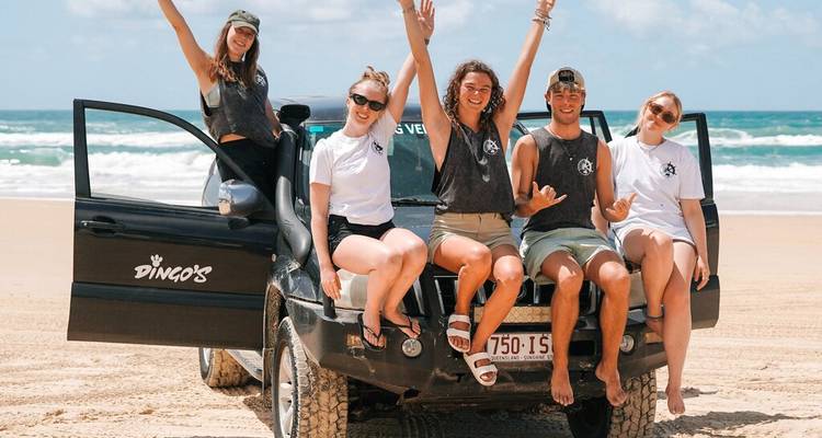 Groupe d'amis posant sur un véhicule 4x4 sur une large plage de sable fin avec des vagues turquoise en arrière-plan.