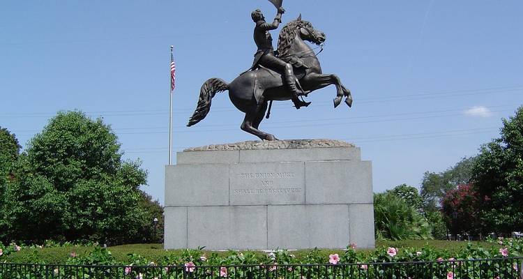 Bronzestatue eines Reiters auf einem aufbäumenden Pferd auf einem Steinsockel, umgeben von Blumen.