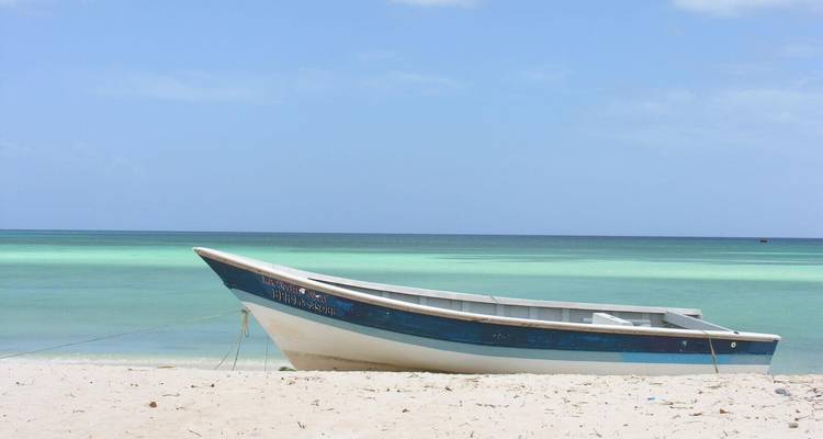 Simple wooden fishing boat rests on white sand against turquoise gradient waters and blue Caribbean sky.