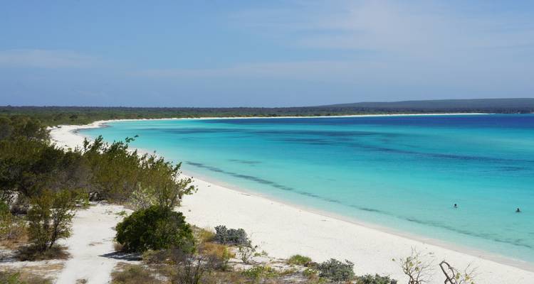 Pristine white-sand beach curving along turquoise water with a distant tree-lined shore under a clear blue sky.