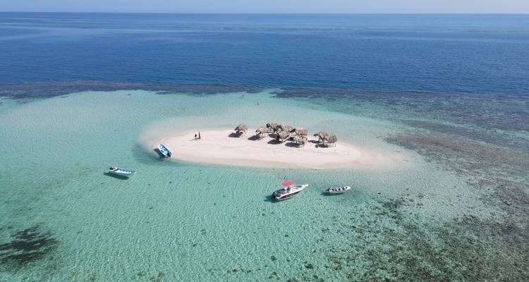 Wide aerial of coral-fringed sandbar island in clear aquamarine sea with small boats anchored nearby.