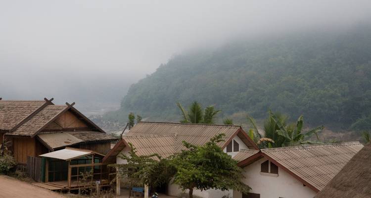 Pequeño pueblito rural con colinas envueltas en niebla detrás de casas de madera y árboles de plátano