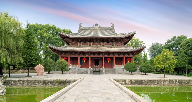 Symmetrische Chinese tempel met aan de voorkant een reflectieënde vijver en omgeven door groen onder een pastelkleurige hemel.