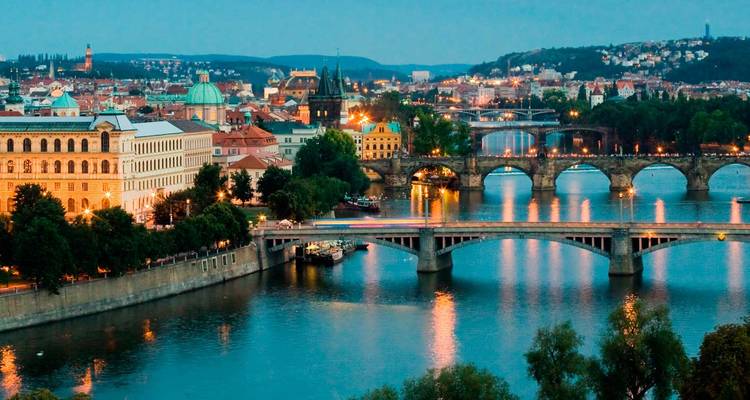 Paisaje urbano nocturno de Praga con el río Vltava y varios puentes de piedra históricos iluminados contra un cielo crepuscular.