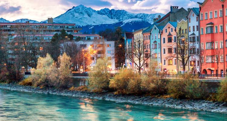 Farbenfrohe Häuser am Flussufer mit schneebedeckten Alpengipfeln, die sich in dem türkisfarbenen Fluss spiegeln.