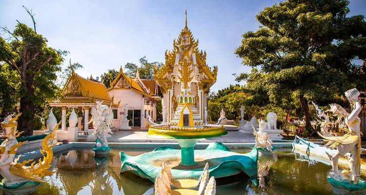 Temple thaïlandais orné avec toit doré situé à côté d'un bassin décoratif rempli de créatures sculptées sous des cieux dégagés.