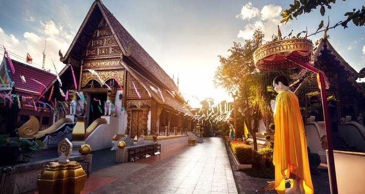 Cour paisible de temple à Chiang Mai illuminée par le soleil couchant, avec une statue de Bouddha vêtue d'une robe dans l'avant-plan.