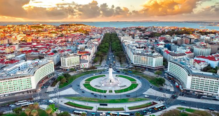 Panorama aéreo de Lisboa que muestra la rotonda del Marqués de Pombal, el bulevar arbolado y el estuario del Tejo bajo nubes dramáticas.