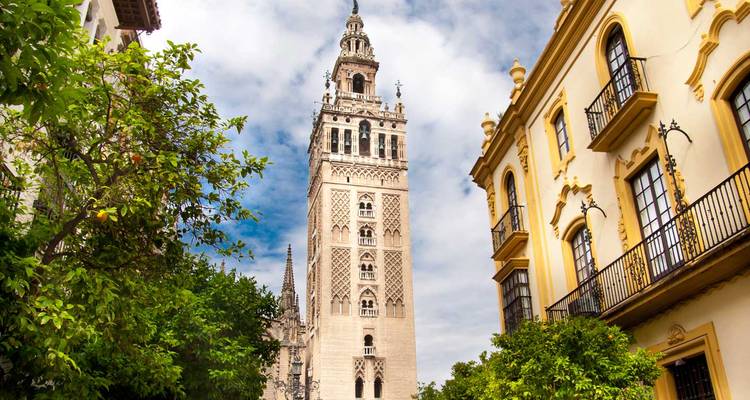 La haute tour Giralda s'élève au-dessus des orangers et des bâtiments historiques sous un ciel partiellement nuageux.