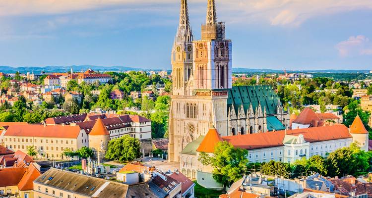 Zagreber Dom mit zwillingsspitzen, die sich über eine grüne Stadtlandschaft und orangefarbene Dächer im weichen Nachmittagslicht erheben.