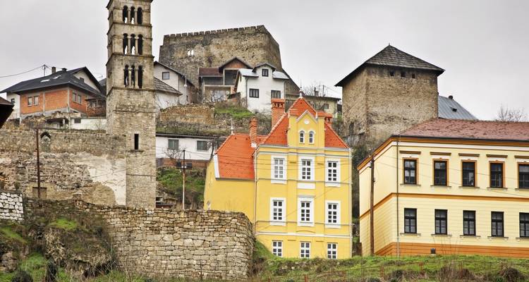 Mittelalterliche Bergstadt mit gelben Häusern und Steintürmen, die sich über alte Mauern unter bewölktem Himmel erheben.