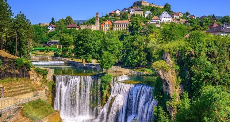 Üppige Landschaft mit dem beeindruckenden mehrstufigen Pliva-Wasserfall von Jajce, darüber die historische Stadt und Festung.