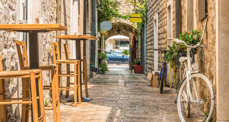 Charmante enge Steingasse, gesäumt mit Holztischen, Pflanzen und einem weißen Fahrrad in einer mediterranen Altstadt.