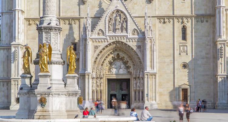 Detail der Fassade der Zagreber Kathedrale mit goldenen Statuen und unscharfen Touristen, die in der Nähe eines Brunnens sitzen.