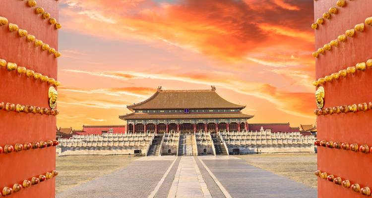 Forbidden City viewed through ornate red palace gates at dramatic sunrise.