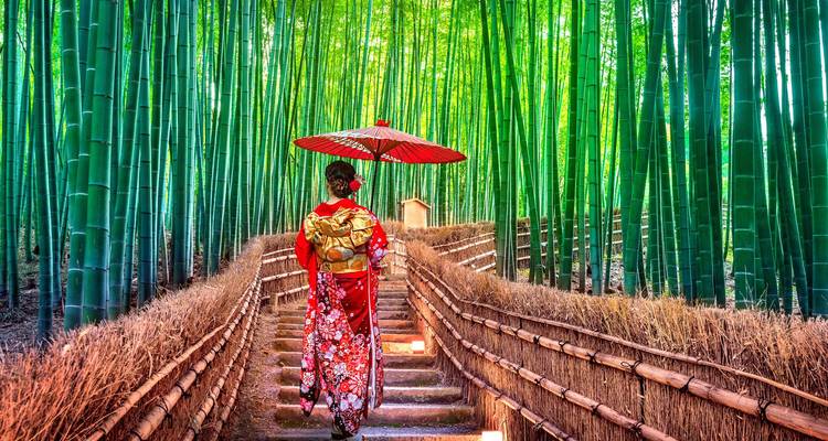 Woman in red kimono with parasol walking up lantern-lit steps through vibrant green bamboo forest.