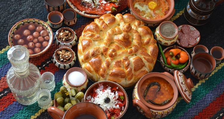 Traditionelle bulgarische Festtafel mit Brot, Salaten, Eingelegtem und Keramikgeschirr auf einem farbenfrohen gewebten Tuch.