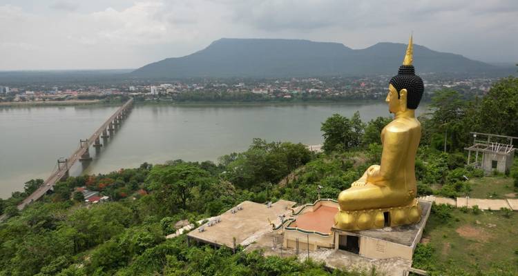Statue de Bouddha dorée au sommet d'une colline face à un long pont enjambant un large fleuve avec une ville lointaine et une chaîne de montagnes en arrière-plan