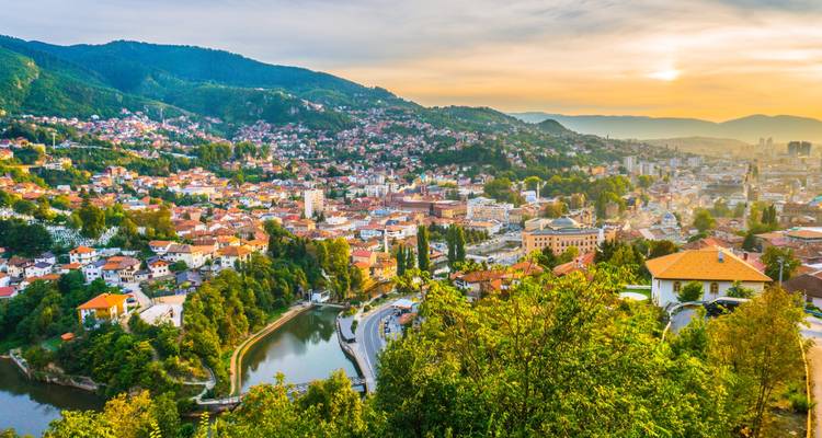 Breiter Panoramablick auf Sarajevo, eingebettet in grüne Hügel mit roten Dächern und Fluss bei Sonnenuntergang.