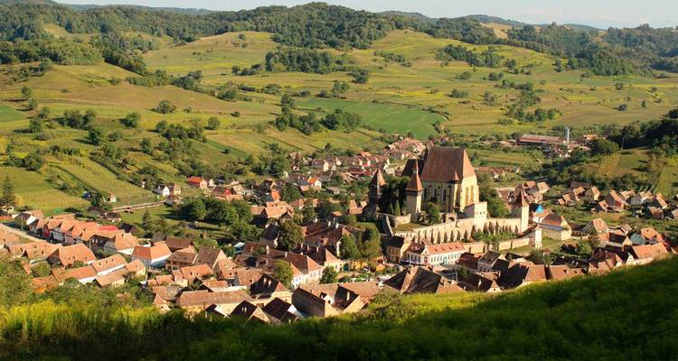 Luftaufnahme eines siebenbürgischen Dorfes, das sich zwischen sanften grünen Hügeln erstreckt, mit einer großen befestigten Kirche im Zentrum.