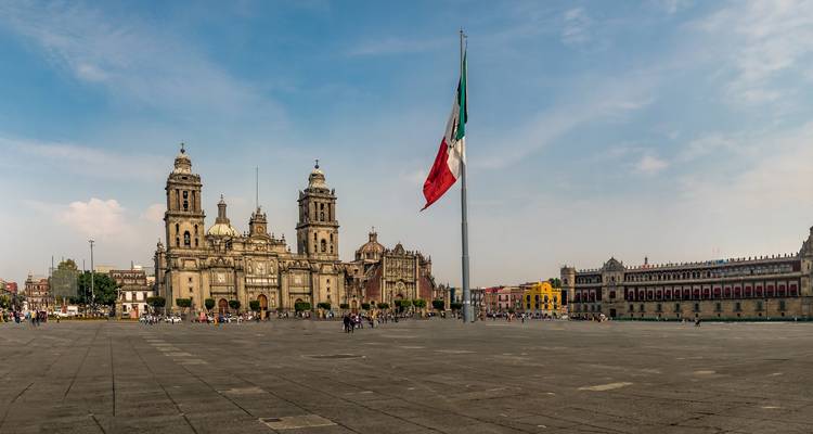Weitläufige Ansicht des Zócalo von Mexiko-Stadt mit der Metropolitankathedrale und einer großen mexikanischen Flagge unter blauem Himmel.