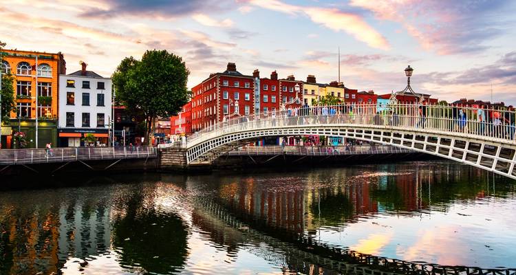 Ha'penny Bridge, das sich bei Sonnenuntergang über den River Liffey in Dublin wölbt, mit farbenfrohen Fassaden, die sich darunter spiegeln.
