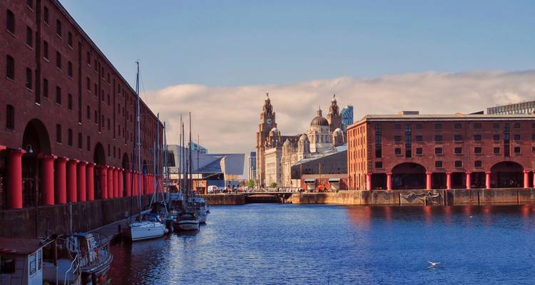 Der Albert Dock Bereich in Liverpool mit historischen Lagerhäusern und der Skyline der Three Graces jenseits einer blauen Uferpromenade.