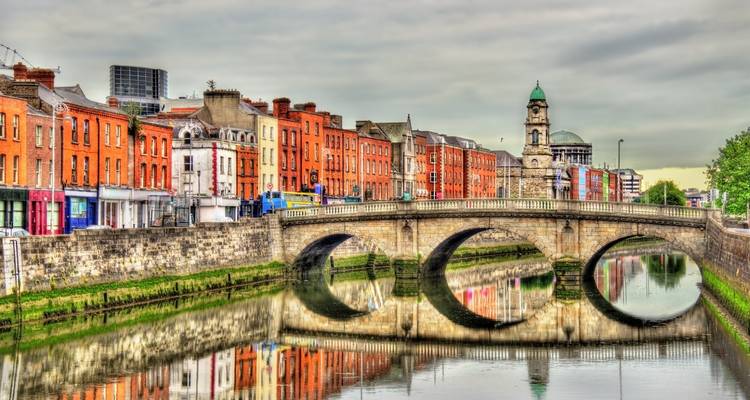 Steinbrücke mit drei Bögen, die den River Liffey überquert und von farbenfrohen historischen Häusern gesäumt wird, unter einem grauen Himmel.