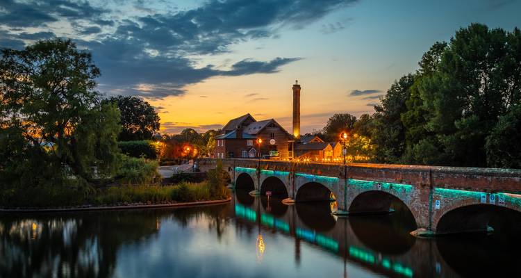 Abendliche Szene von Stratford-upon-Avon mit einer gemauerten Bogenbruecke und einer Fluessmuehle am Fluss, die sich in ruhigem Wasser spiegeln.