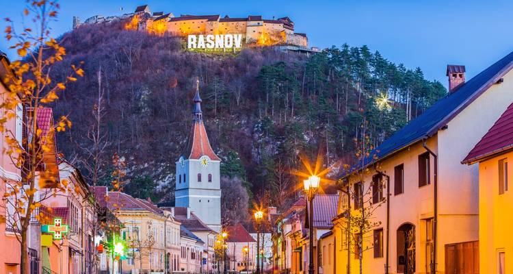 Twilight street view toward Rasnov Fortress sign lit up on the hillside above the town.