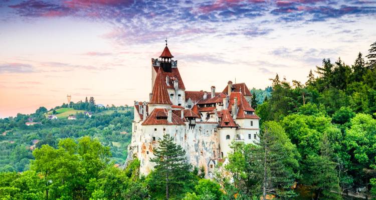 Das märchenhafte Schloss Bran thront auf einem bewaldeten Hügel vor einem farbenfrohen Sonnenuntergangshimmel in Transsilvanien.