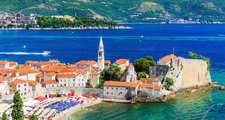 Die Altstadt von Budva und der Strand mit dem leuchtend blauen Adriatischen Meer und entfernten Hügeln im Hintergrund.