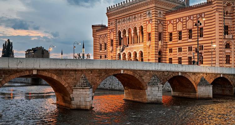 Das prächtig verzierte Rathaus von Sarajevo erleuchtet den Abendhimmel über dem Fluss Miljacka und der historischen Brücke.