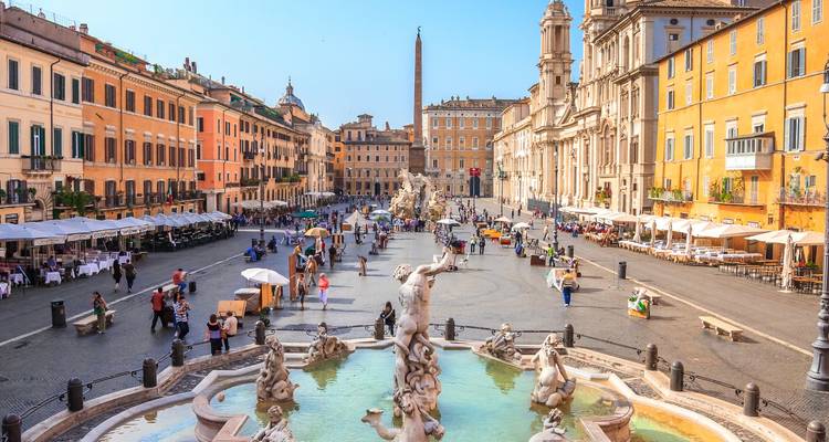 La Piazza Navona animée à Rome avec des parasols de café, des fontaines baroques et les bâtiments ocre environnants sous un ciel bleu éclatant.