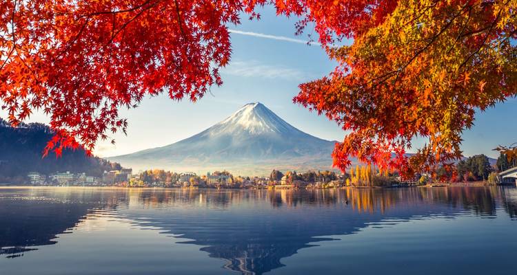 Iconische Mount Fuji met sneeuw bedekt weerspiegeld in het rustige Lake Kawaguchiko omringd door levendige rode herfstbladeren van esdoorns bij zonsopgang.