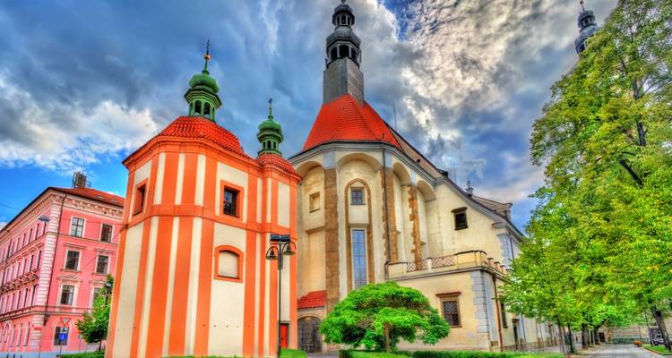 Iglesia barroca vibrante con fachada naranja rayada y cúpulas verdes al lado de una torre más alta con techo rojo bajo nubes dramáticas.