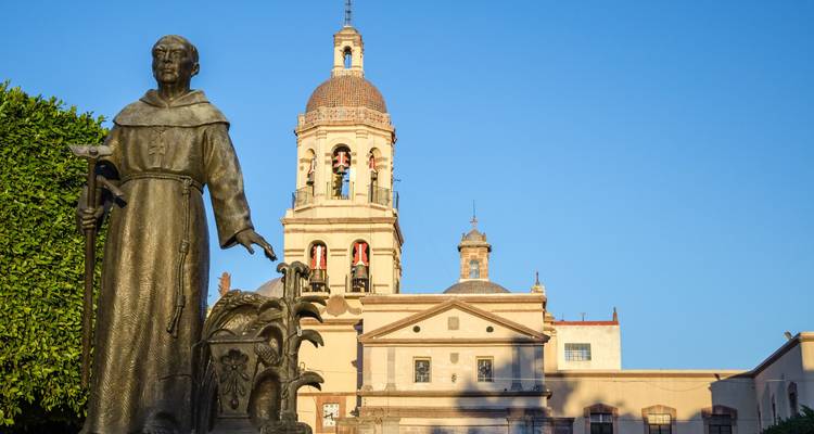 Statue eines franziskanischen Mönchs vor einer historischen Kirche mit Glockenturm unter klarem blauen Himmel