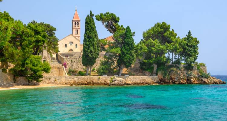 Kleine Steinkirche und Zypressenbäume an einem abgelegenen adriatischen Strand mit klarem türkisfarbenem Wasser.