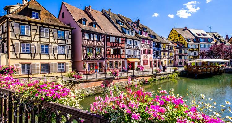 Colorful half-timbered houses with flower boxes line a canal in a picturesque old town under a blue sky.