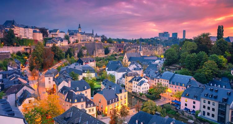 Twilight panorama of a fortified European city with dramatic cliffs and church spires above cozy homes.
