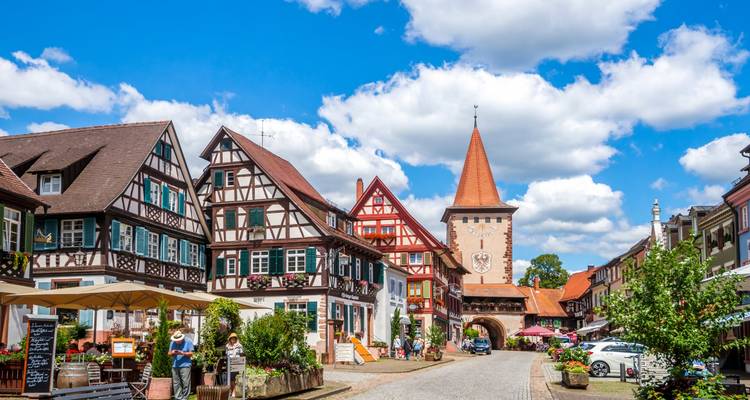 Quaint German village street with timber-framed houses leading to a tall gate tower under blue skies.