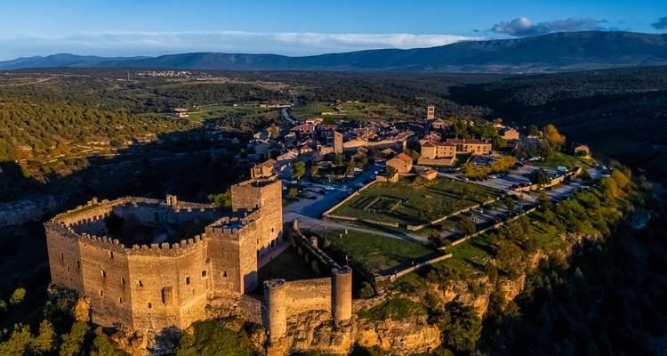 Vue aérienne d'un château médiéval perché sur une colline et d'un petit village entourés de paysages forestiers au lever du soleil.