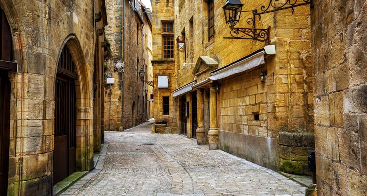 Ruelle étroite pavée de galets avec des murs de pierre médiévale, des lampadaires en fer forgé et des fenêtres à volets dans la vieille ville de Sarlat.